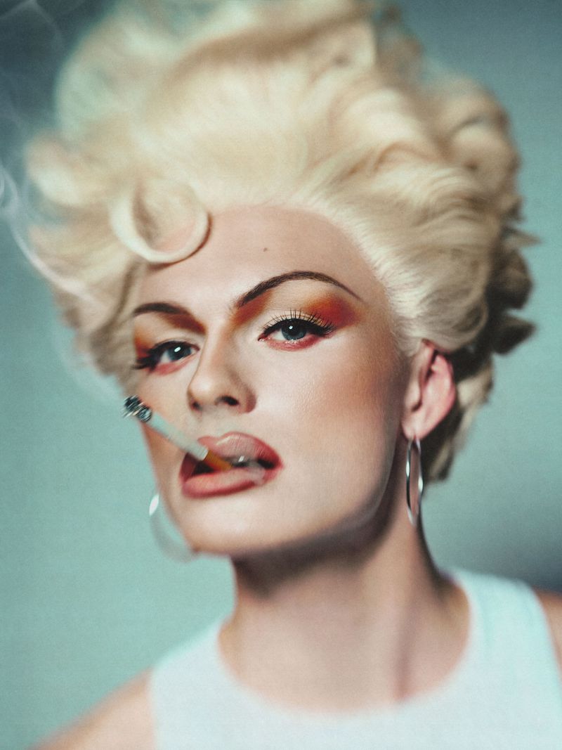 Portrait of a person with platinum blonde hair, theatrical makeup, and large hoop earrings smoking a cigarette against a pale blue background.