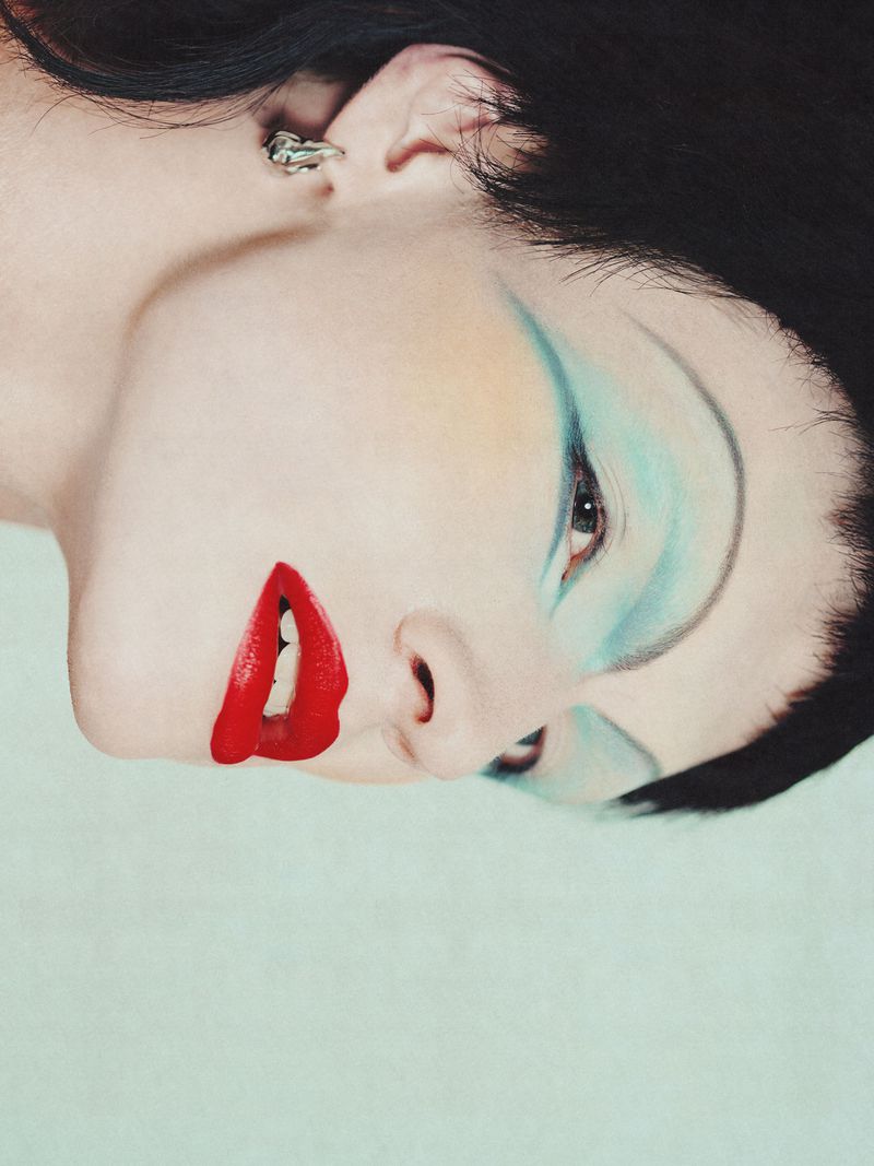 Close-up portrait of a woman with dramatic makeup featuring bright red lipstick and blue eyeshadow, shot against a solid light blue background.