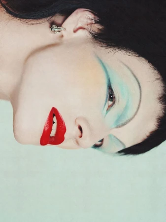 Close-up portrait of a woman with dramatic makeup featuring bright red lipstick and blue eyeshadow, shot against a solid light blue background.