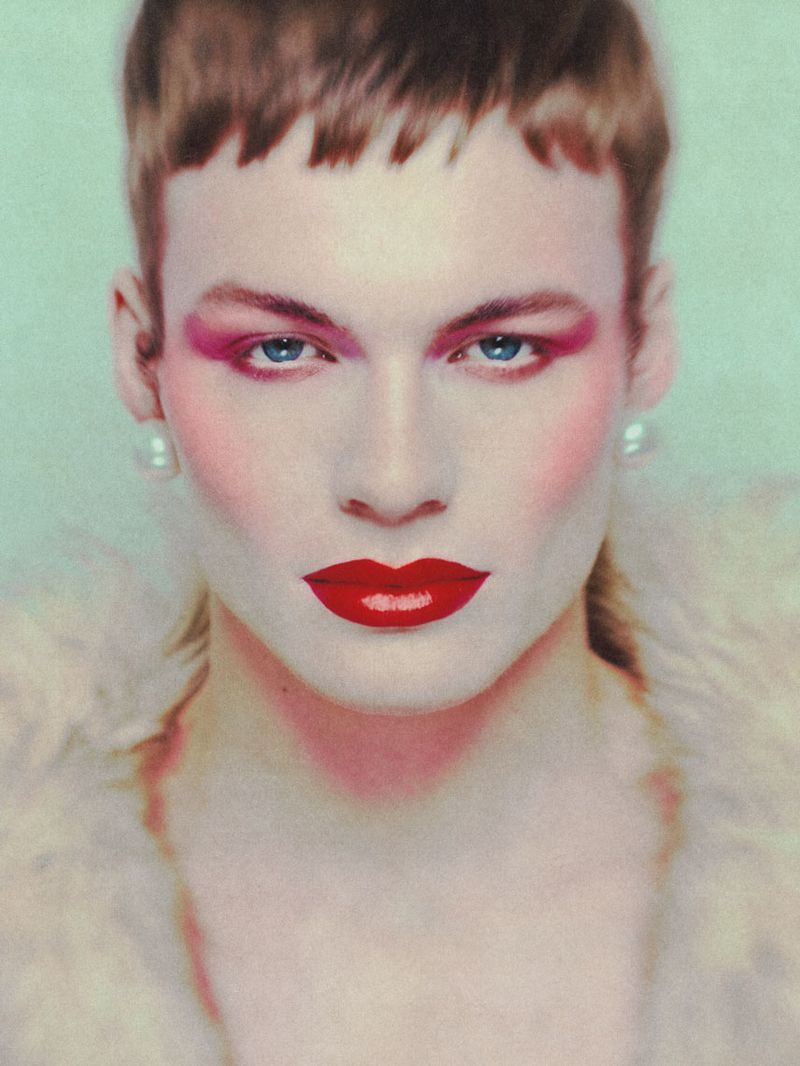Close-up portrait of an adult with light skin, red lipstick, and pink eyeshadow, wearing pearl earrings and a fur shawl against a pale green background.