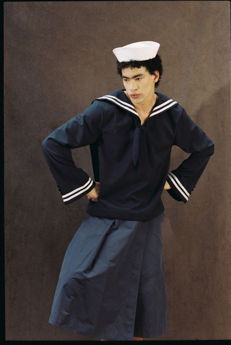 Person in navy sailor uniform and white hat standing against a brown background, posing with hands on hips, looking off to the side.
