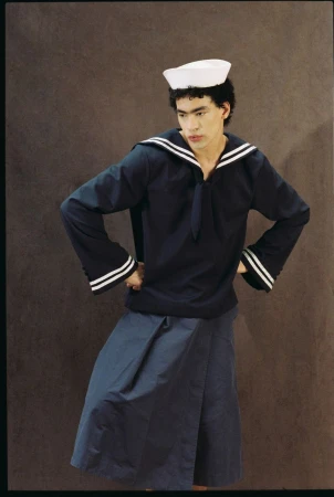 Person in navy sailor uniform and white hat standing against a brown background, posing with hands on hips, looking off to the side.