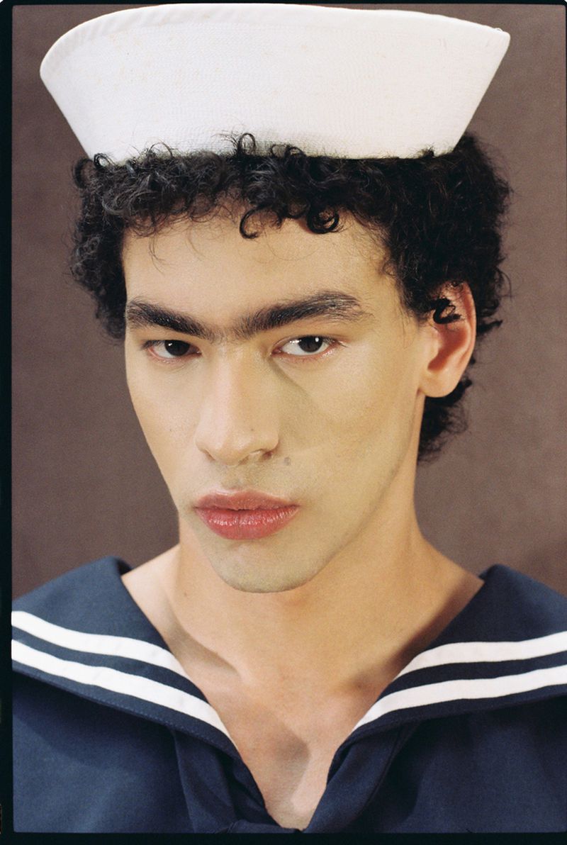 Close-up studio portrait of a young adult wearing a sailor hat and navy and white shirt against a brown background.