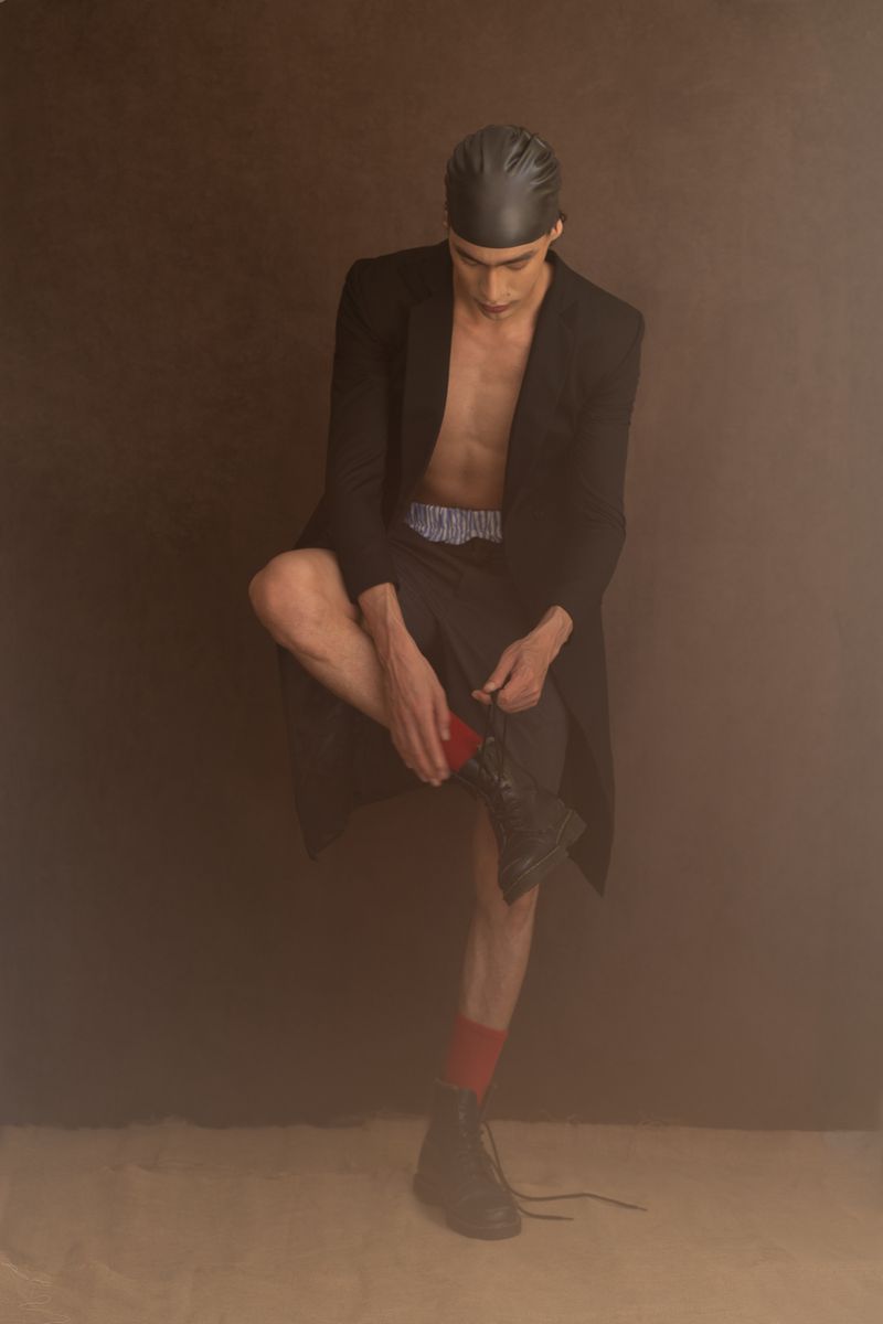 Man in black coat and head covering, seated and putting on boot, in a studio with brown background and neutral-tone floor.