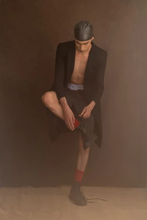 Man in black coat and head covering, seated and putting on boot, in a studio with brown background and neutral-tone floor.