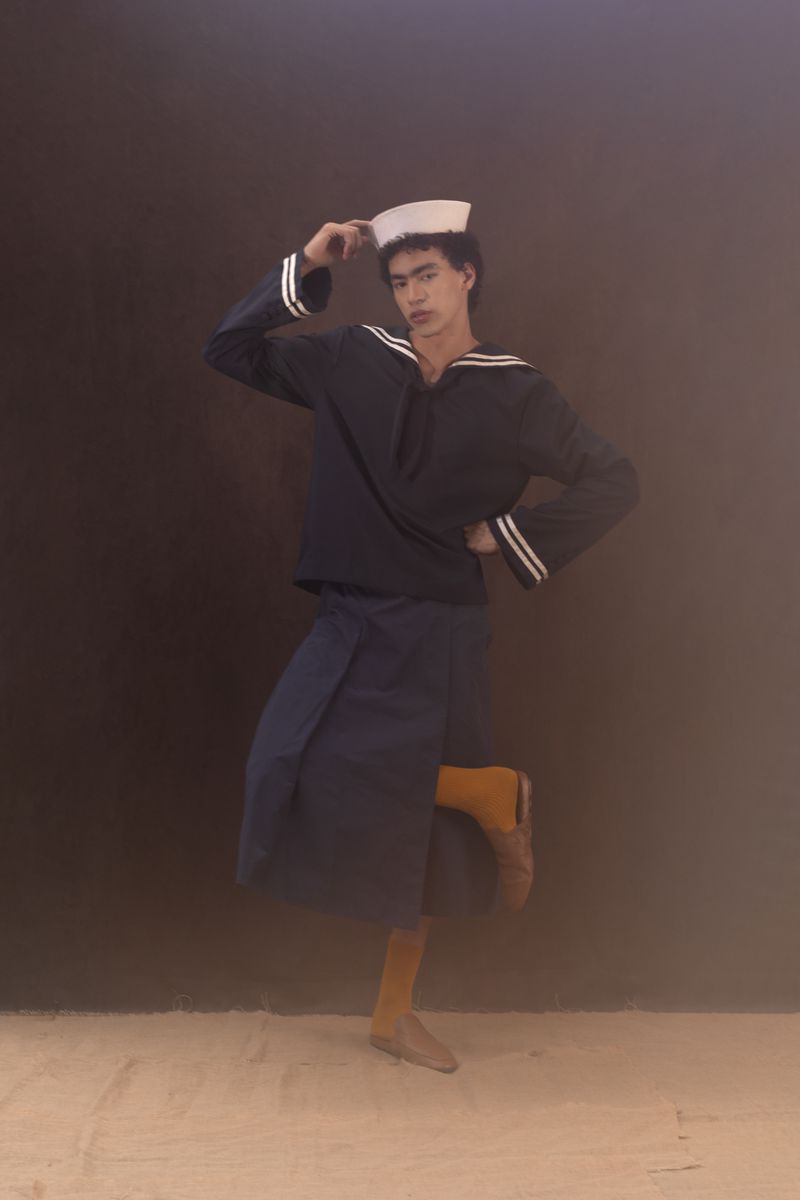 A young person in a navy sailor outfit poses in front of a dark background, wearing a sailor hat, blue top, and a wrapped skirt.