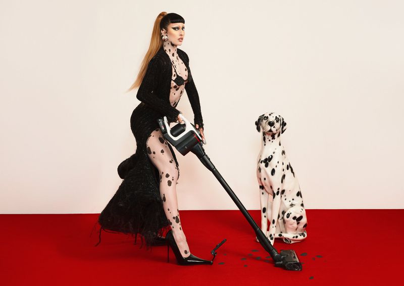 Glamorous woman vacuums in designer dress and heels with a Dalmatian sitting to the side, set against a bright red floor.