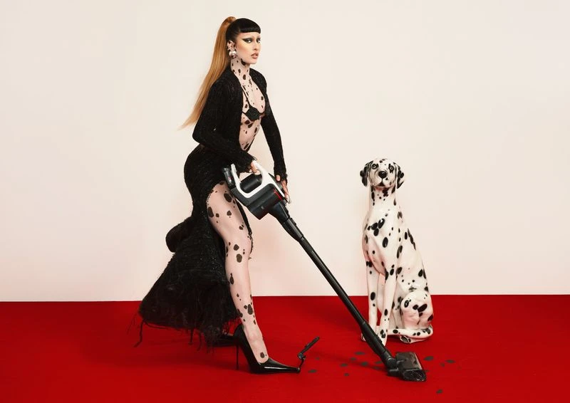 Glamorous woman vacuums in designer dress and heels with a Dalmatian sitting to the side, set against a bright red floor.