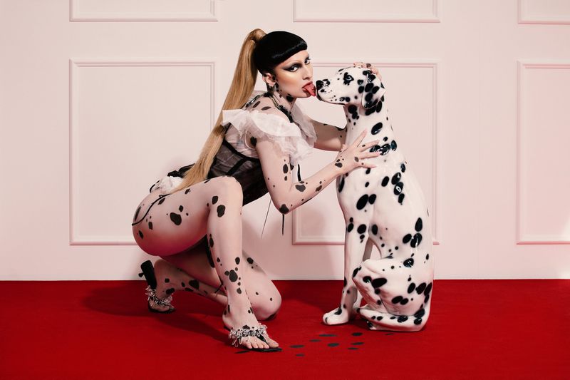 Woman in Dalmatian-print bodysuit poses with a Dalmatian dog on red carpet in front of white paneled wall, eye-level angle.