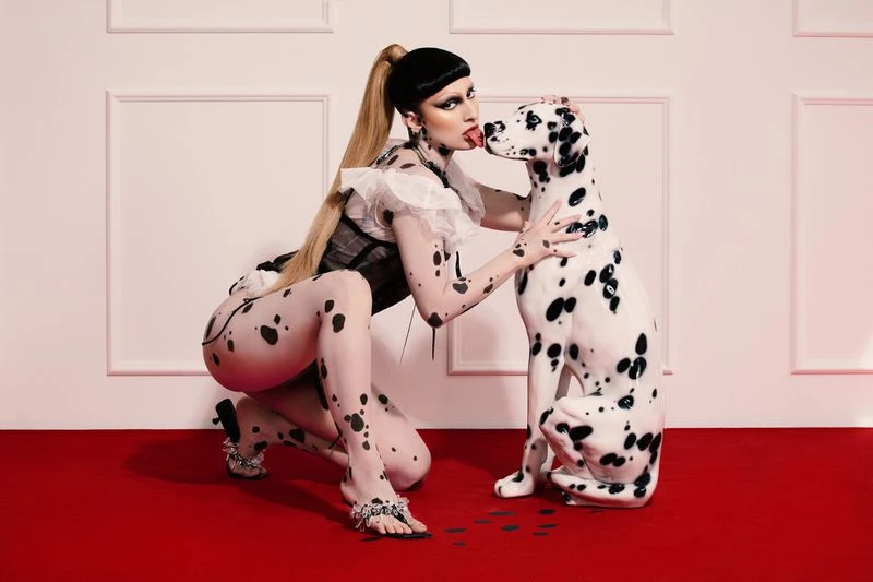 Woman in Dalmatian-print bodysuit poses with a Dalmatian dog on red carpet in front of white paneled wall, eye-level angle.
