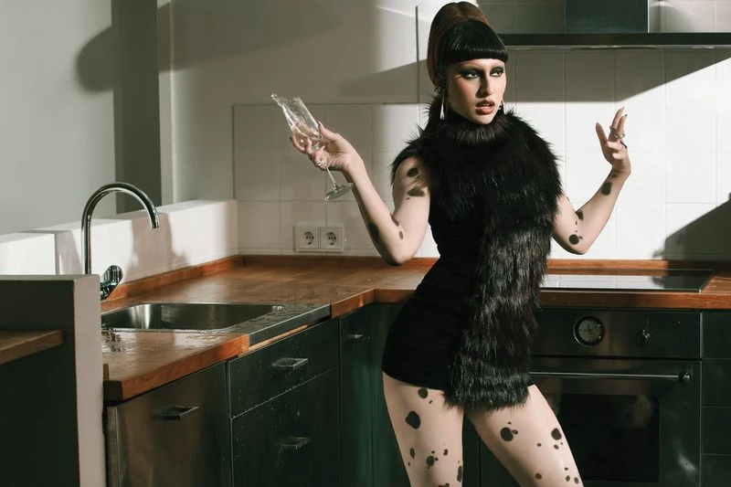 Stylish woman in a black dress with spotted tights holds a wine glass in a modern kitchen with dark cabinets and a stainless steel sink.