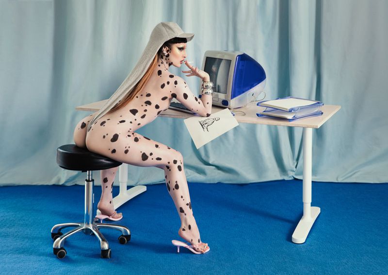 Woman in body paint sits at desk with computer. Nude with dalmatian pattern, wearing hat, working from home in blue office setting.