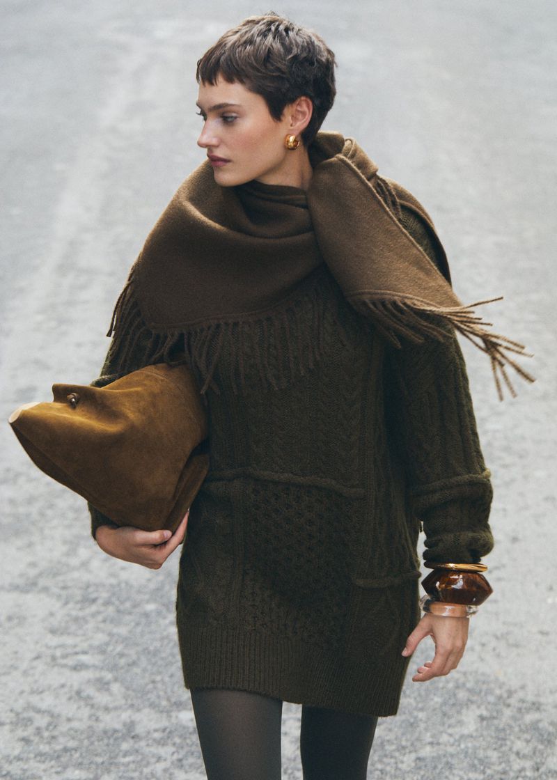 Woman in an olive green knit dress and scarf holds a suede bag against a grey background in a fashion editorial-style portrait.
