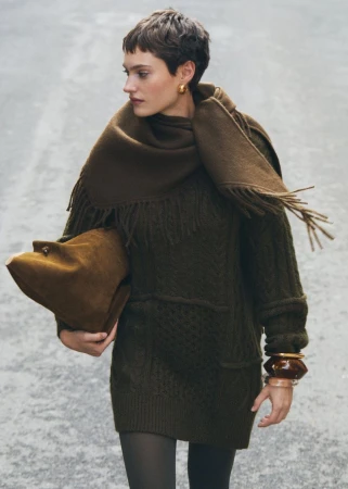 Woman in an olive green knit dress and scarf holds a suede bag against a grey background in a fashion editorial-style portrait.