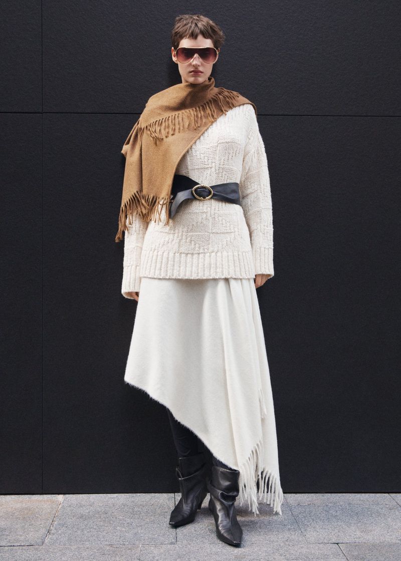Adult female fashion model wearing a cream-colored knit dress with a brown fringed scarf and black leather boots against a black wall.