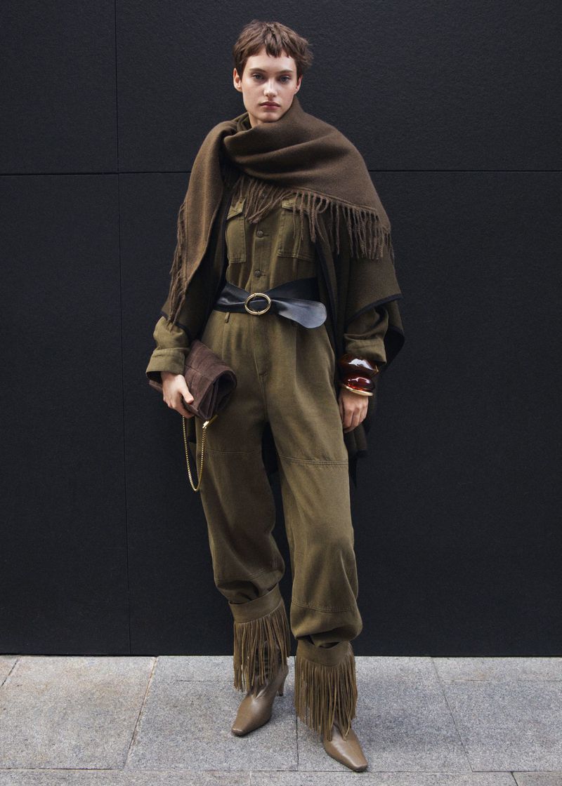 A young woman in a green fringe-trimmed jumpsuit with a brown fringed shawl stands against a dark gray wall holding a brown clutch.