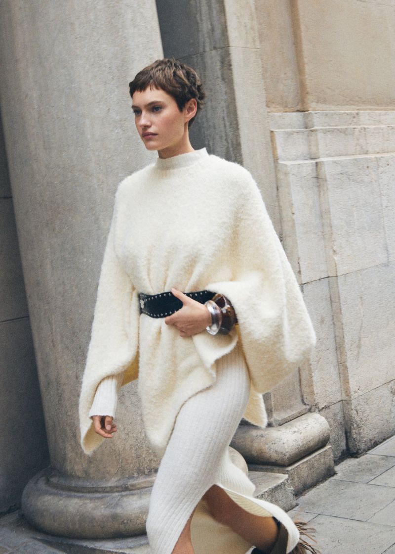 Woman in a cream-colored sweater dress and wide belt posing in front of a building with stone columns in an outdoor setting.