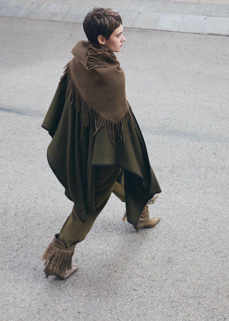 Woman in olive green cape and fringed boots walks on asphalt.  Fashion editorial shot from above. Minimalist aesthetic.