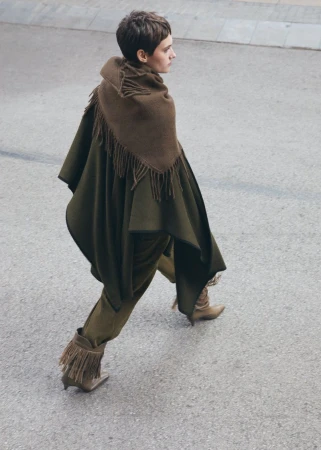 Woman in olive green cape and fringed boots walks on asphalt.  Fashion editorial shot from above. Minimalist aesthetic.