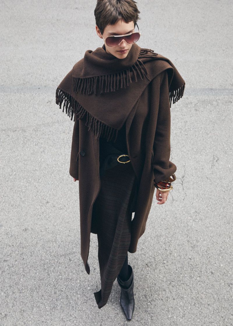 A stylish adult woman in a long brown coat, fringed scarf, and sunglasses poses on an asphalt surface in an eye-level outdoor shot.