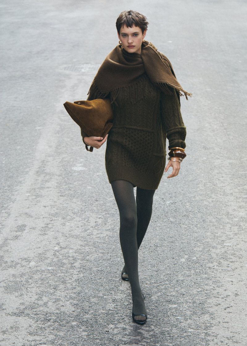 Adult female wearing a brown sweater dress, tights, and heels walks on an urban street with a fringed scarf and handbag.