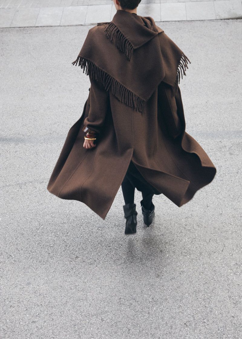 A person in a brown fringe coat walks on a grey concrete surface, showcasing a stylish and unique fashion statement with a flowing silhouette.