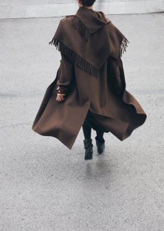 A person in a brown fringe coat walks on a grey concrete surface, showcasing a stylish and unique fashion statement with a flowing silhouette.