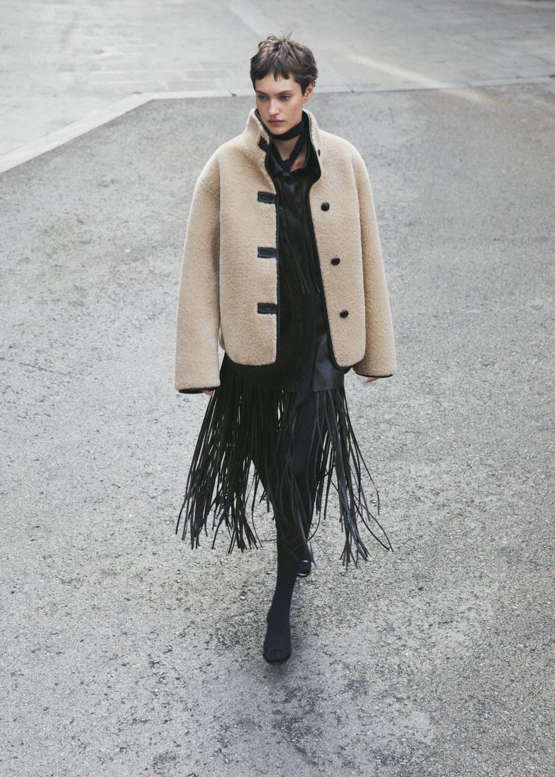 A woman in a shearling jacket and a skirt with long fringe, shot from above on a textured grey surface, fashion-forward attire and pose.