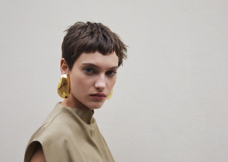 Portrait of an adult female with short brown hair wearing oversized gold earrings and a tan top, posing in front of a plain wall.
