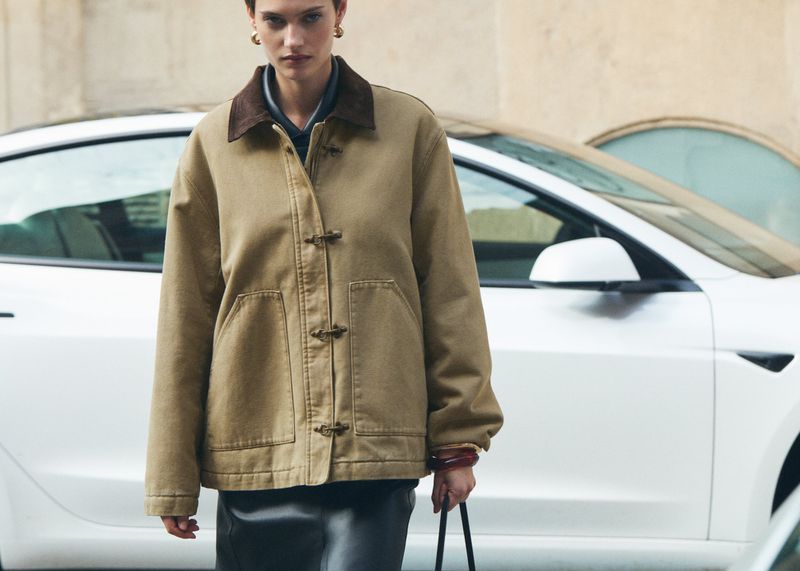 Model wearing a tan jacket with toggle fastenings standing outdoors in front of a white car
