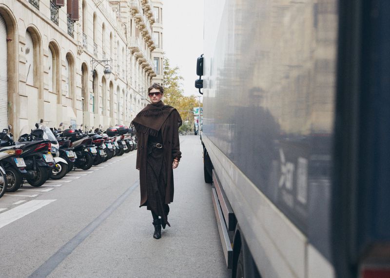 A woman wearing a brown coat and black boots walks down a city street next to parked scooters and a truck on a cloudy day.