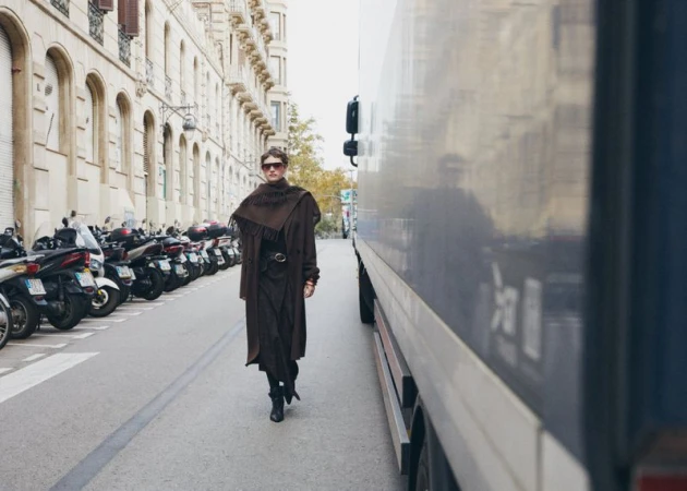 A woman wearing a brown coat and black boots walks down a city street next to parked scooters and a truck on a cloudy day.