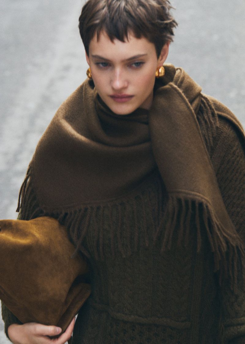 A woman poses with an olive green fringed scarf and a brown handbag, her gaze cast downward, and she wears gold earrings.