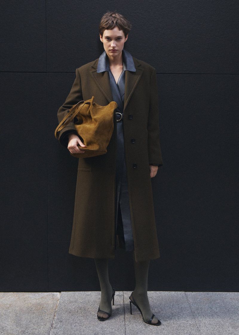 A woman in a dark coat poses against a black wall, carrying a brown bag and wearing sheer dark hose and open-toed heels in a fashion shot.