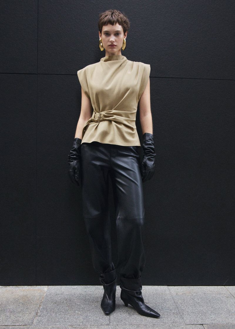 Woman in tan sleeveless top, black leather pants, and gloves poses against a dark grey wall wearing hoop earrings and ankle boots.