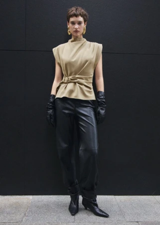 Woman in tan sleeveless top, black leather pants, and gloves poses against a dark grey wall wearing hoop earrings and ankle boots.