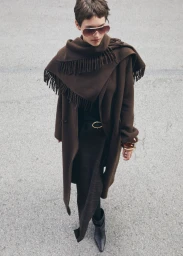 Model wearing a brown coat and fringe scarf with tinted sunglasses standing on textured pavement for Mango Wild Spirit