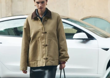 Model wearing a tan jacket with toggle fastenings standing outdoors in front of a white car