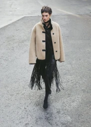 Model wearing a textured cream jacket and black fringed skirt walking outdoors on pavement.