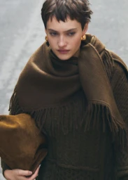Model with short hair wearing an olive green sweater, matching scarf, and holding a brown clutch against a blurred outdoor background