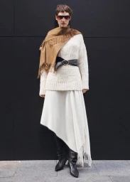Model wearing a white sweater, cream skirt, brown fringed scarf, and dark sunglasses against a dark wall