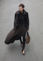 Model in dark faux fur jacket and long pleated skirt walking outdoors, holding a brown shoulder bag.