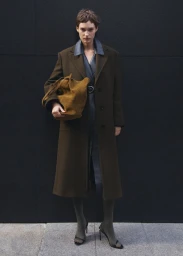 Model standing outdoors wearing a long brown coat and holding a brown suede handbag against a dark wall