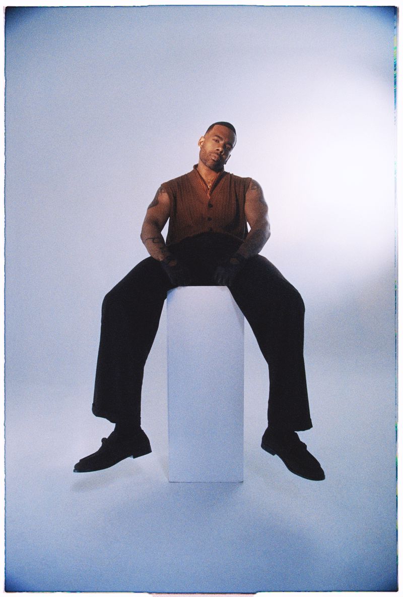 Adult male with tattoos seated on a white pillar in photo studio with brown shirt, black pants, and black dress shoes against gray backdrop.