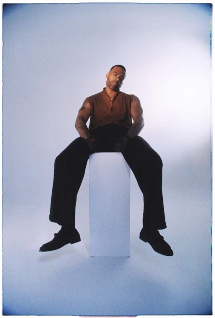 Adult male with tattoos seated on a white pillar in photo studio with brown shirt, black pants, and black dress shoes against gray backdrop.