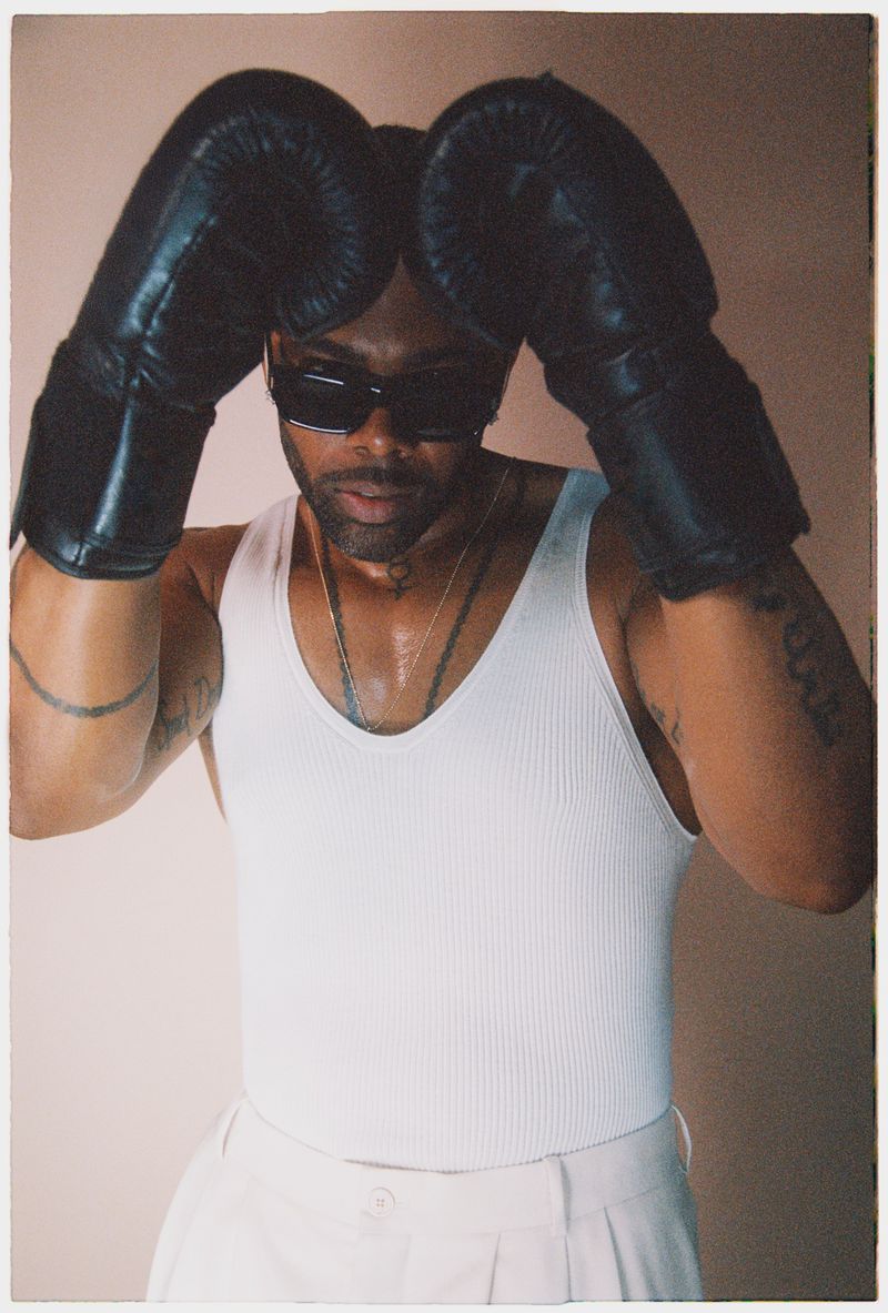 Portrait of an African American male wearing boxing gloves, sunglasses, and a white tank top against a neutral background.