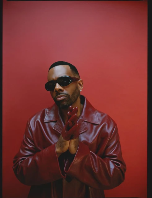 Portrait of a Black man wearing sunglasses and a red leather jacket against a red background, hands clasped together in a neutral studio setting.