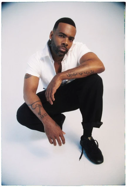 Portrait of an African American man with tattoos, wearing a white shirt and black pants, squatting in a photo studio with a grey background.