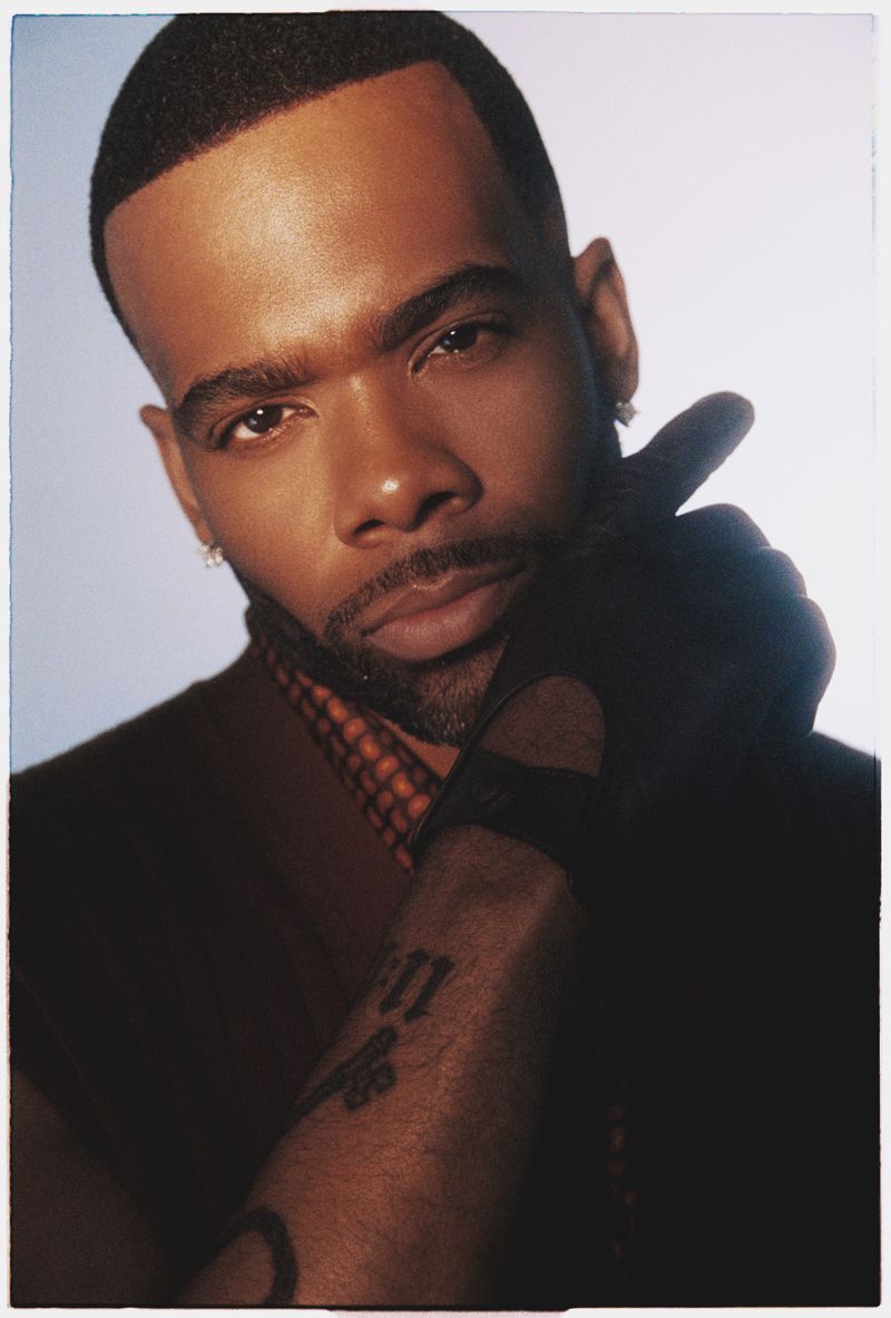 Portrait of an African American man with facial hair and a hand raised to his face wearing a brown vest and black glove in a studio.