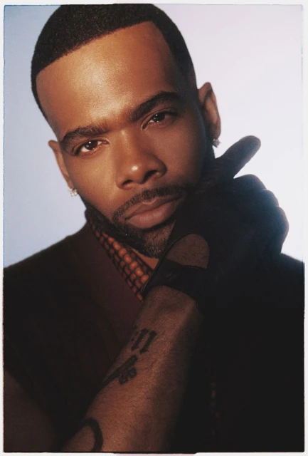Portrait of an African American man with facial hair and a hand raised to his face wearing a brown vest and black glove in a studio.